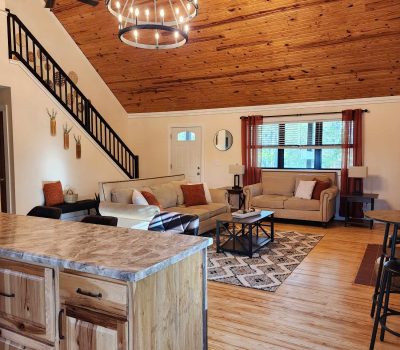 A spacious living room with a high, wooden vaulted ceiling and a large chandelier. The room features two beige sofas with orange pillows, a patterned rug, and a wooden coffee table. A staircase with black railings leads to an upper level. A kitchen island is visible.