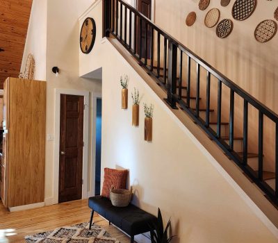 A modern hallway with a wooden ceiling and light-colored walls. It features a black bench topped with cushions, a patterned rug, and a potted plant. Stairs with a black railing lead up, with decorative baskets adorning the wall above.