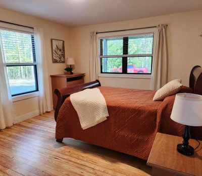 A cozy bedroom with a queen-sized bed covered in a rust-colored quilt. The room features wood flooring, two large windows with white curtains, a small wooden nightstand with a lamp, a wooden dresser with a lamp and decor, and a painting on the wall.