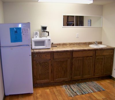A small kitchen area features a white refrigerator with a blue flyer taped to it, a microwave, a coffee maker, wooden cabinets, a sink, and a small mirror above the counter. The floor is wooden with a throw rug, and a trash can is situated to the right of the cabinets.