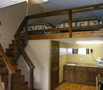 A room with a wooden loft bed placed above a kitchenette. The loft is accessed by wooden stairs on the left. The kitchenette includes a sink, microwave, and coffee maker, with cabinets below and a mirror above. The room has wooden flooring and beige walls.