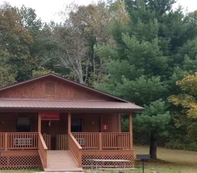A small wooden cabin with two units labeled "1" and "2" sits amidst a forested area. The cabin has a front porch, central steps, and a wooden railing. Tall trees, including evergreens, surround the cabin, creating a serene backdrop. A grill is near the cabin.