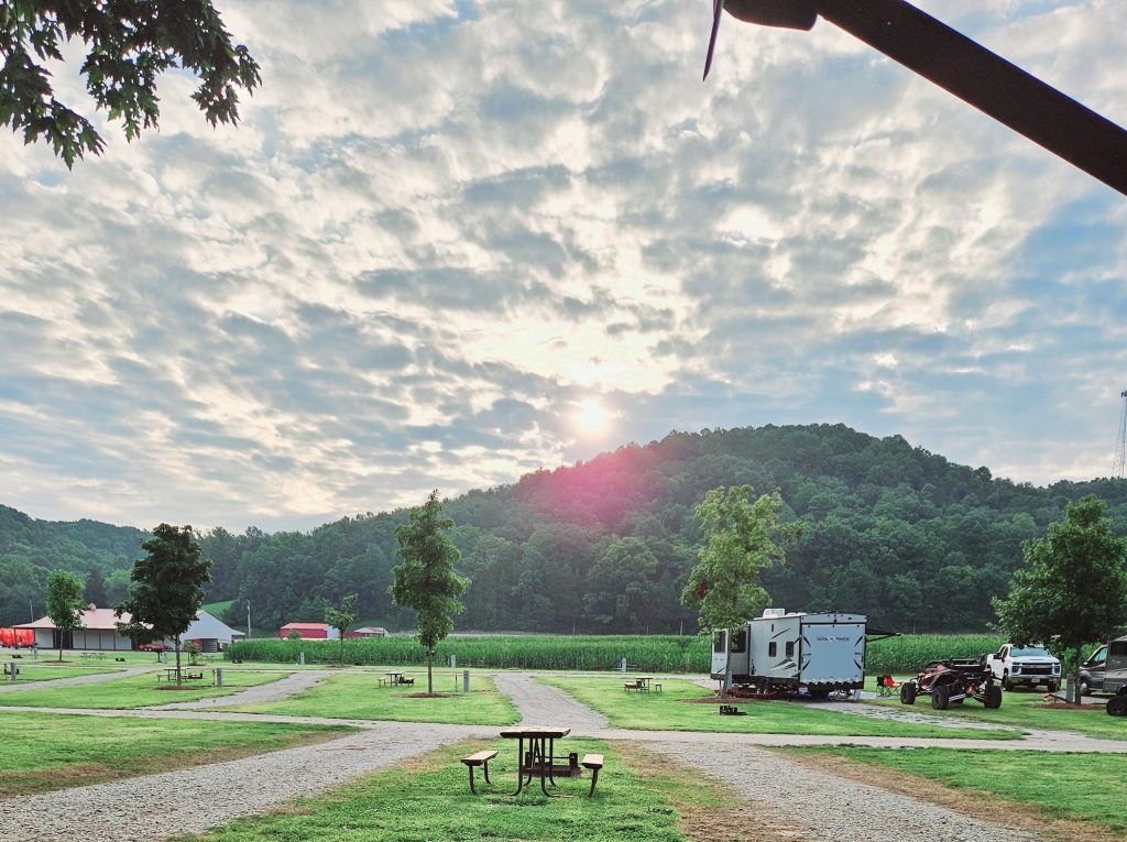 A scenic view of a campsite at sunrise, featuring a camper van parked on grassy terrain. Picnic tables are scattered around, with trees and a distant hill covered in foliage. Partly cloudy sky is illuminated by the sun.