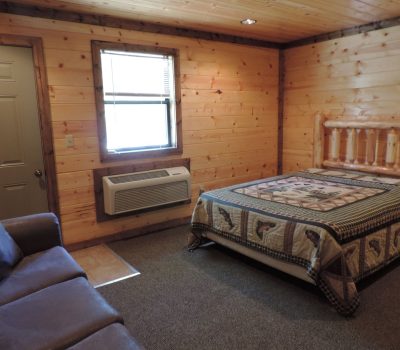 A cozy log cabin room with wooden walls and ceiling. The room features a bed with a patchwork quilt, a brown sofa, a window air conditioning unit, and a green door. A window with blinds lets in natural light. The floor has carpeting and tile near the door.