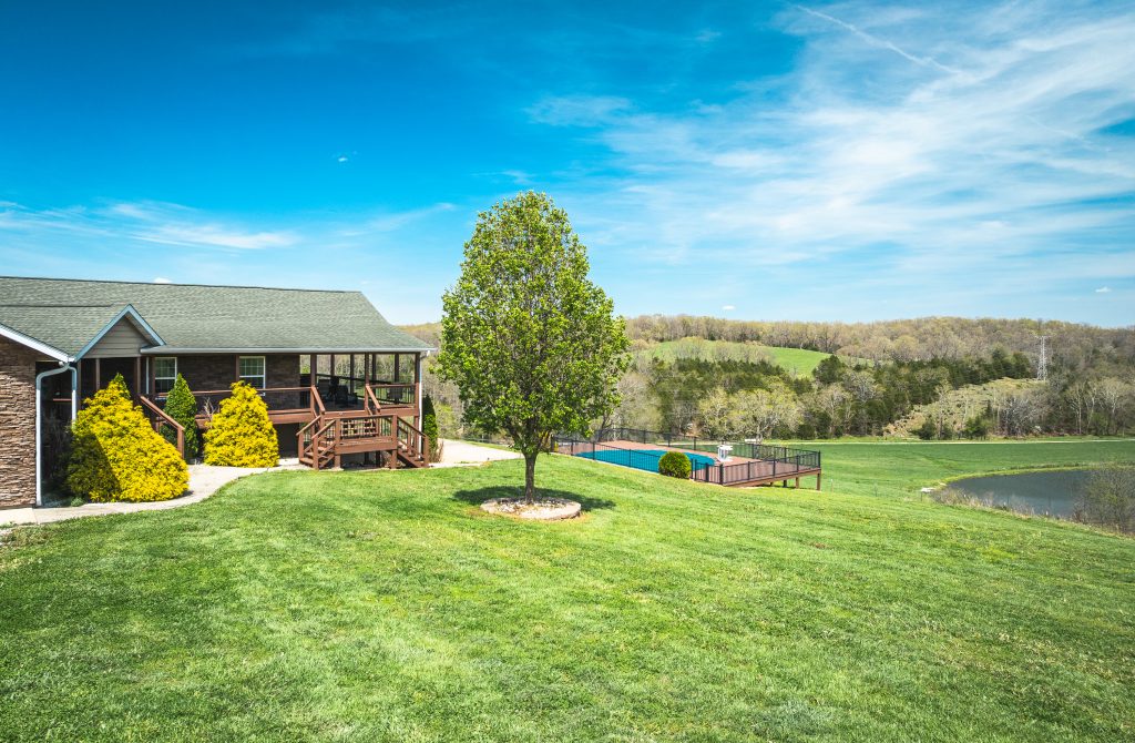 A house with a porch sits on a large, green lawn. A tree stands nearby, and there is a fenced swimming pool in the distance. The landscape features rolling hills and a blue sky with wispy clouds.