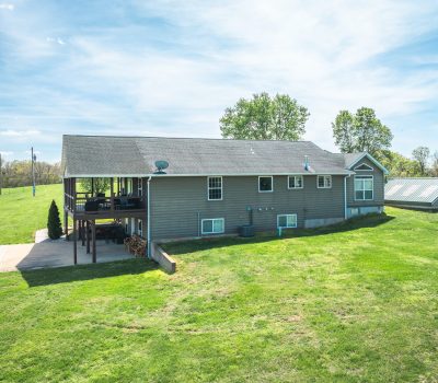 A gray house with a deck stands on a large, green grassy lawn with trees in the background under a blue sky. A small outbuilding with a metal roof is nearby.