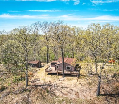 A small cabin with a wooden deck and separate shed is surrounded by tall trees on a sunny day, with a dirt clearing and a partially wooded landscape visible under a bright blue sky.