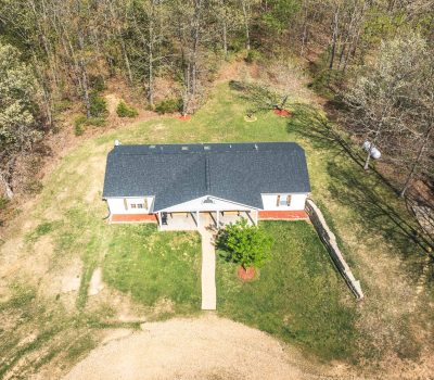 Aerial view of a small white house with a dark roof, surrounded by grass and trees. A walkway leads to the front porch, and the yard has some bare patches and planted shrubs.