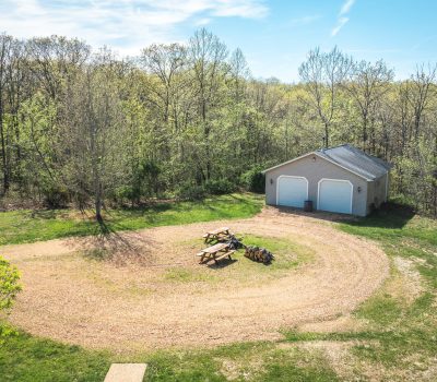 Aerial view of a gravel driveway curving around picnic tables and a small fire pit, leading to a garage with two white doors, surrounded by trees and greenery on a sunny day.