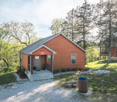 A small wooden cabin with a red exterior sits among trees, with a porch, grill, and a gravel walkway. A trash can is in the grass nearby, and another building is partially visible in the background.