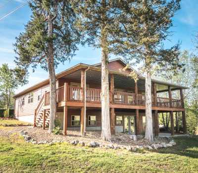 A two-story wooden house with a large wraparound porch, surrounded by tall trees and greenery, sits on a grassy lawn under a clear blue sky.