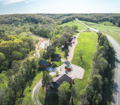 Aerial view of a rural landscape with a winding road, a few houses with red roofs, trees, grassy fields, and a stream running through a wooded area under a clear sky.