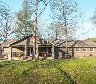 A single-story house with beige siding and a front porch, surrounded by trees and grassy lawn on a sunny day. The home has a gabled roof and sits in a natural, wooded setting.