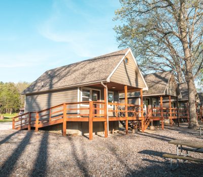 A small wooden cabin with a ramp and porch sits among tall trees under a clear blue sky. Picnic tables and gravel are in the foreground, and a second similar cabin is visible behind it.