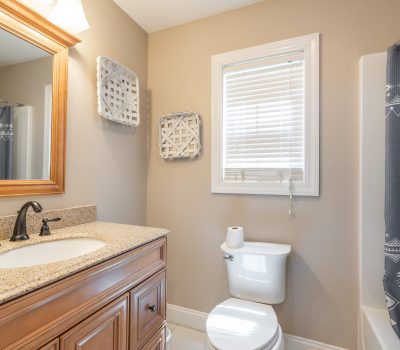 A bathroom with a wooden vanity and granite countertop, a black faucet, a toilet with a toilet paper roll, a window with blinds, wall decor, and a shower area with a patterned gray curtain.