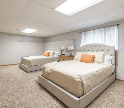 A spacious bedroom with two beige upholstered beds, orange and white accent pillows, a nightstand with lamps between them, beige carpet, neutral walls, and a window with white curtains letting in natural light.