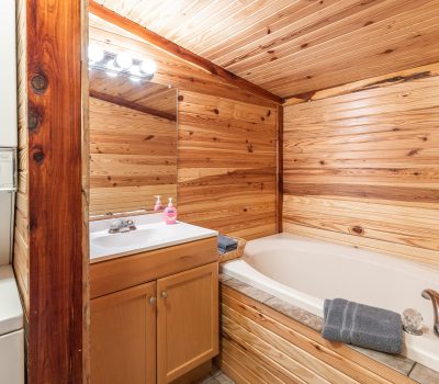 A cozy bathroom with wooden walls and ceiling, featuring a built-in bathtub, a small vanity with a sink, a mirror above, and a stacked washer and dryer unit on the left. Towels and soap are neatly placed.