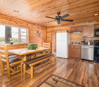 A cozy cabin kitchen with wood-paneled walls and ceiling, a dining table with benches, white refrigerator, black stove and microwave, and large window showing a scenic outdoor view.