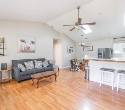 Open-concept living room and kitchen with wood flooring, gray sofas, a coffee table, wall art of a cow, bar stools, ceiling fan, and dining area with table and chairs. Bright natural light from skylights.