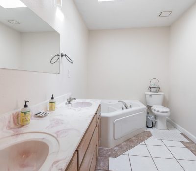 A bathroom with a double sink vanity, a large mirror, a corner bathtub with handrails, and a toilet. The room has light-colored walls and tile flooring with a skylight above.