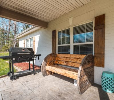 A wooden bench and a black grill sit on a stone patio outside a white brick house with large windows and wooden shutters. Green grass and trees are visible in the background.