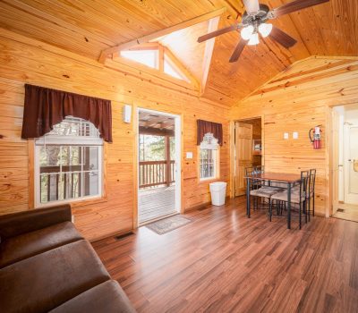 A cozy wooden cabin interior with pine walls and ceiling, ceiling fan, brown sofa, small dining table with four chairs, and large windows with dark curtains overlooking a covered porch.
