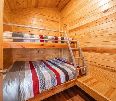 A cozy wooden room with bunk beds; the lower bed is a double and the upper bed is a twin. Both beds have striped bedding in red, gray, and white. A metal ladder leads to the top bunk.