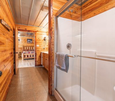 A bathroom with wood-paneled walls, a glass shower with a towel on the handle, brown tile floor, vanity with sink, and a view into a bedroom with a wooden bed in the background.