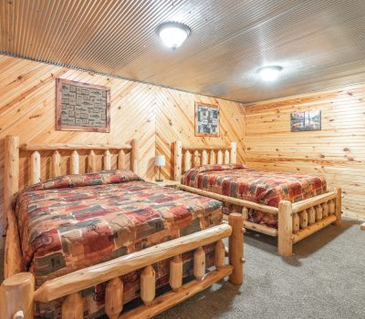 A cozy bedroom with wood-paneled walls and ceiling, featuring two rustic log-frame beds with patterned bedspreads, carpeted floor, wall art, and a small wooden chest at the foot of one bed.