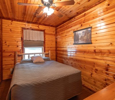 A small cozy bedroom with wood-paneled walls and ceiling, a ceiling fan with lights, a single bed with a gray quilt, a window with a shade, and a nature photo hanging on the wall.