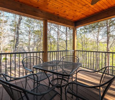 A covered wooden porch with a metal table and four chairs, surrounded by a black railing. Trees and greenery are visible in the background through the screened enclosure.