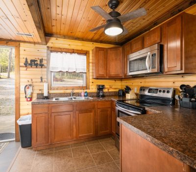 Cozy cabin kitchen with wooden walls and ceiling, brown cabinets, dark countertops, stainless steel appliances, and a window above the sink. A door to the left opens to a view of trees and rocks outside.