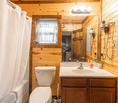 A rustic bathroom with wood-paneled walls, a white shower curtain, toilet, and sink with a mirror above. There are wall-mounted lights above the mirror and a window with a sheer curtain above the toilet.