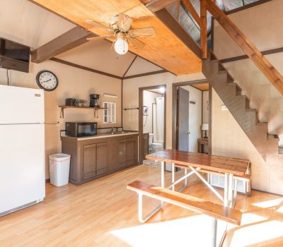 A bright, rustic kitchen with wood paneling, a white refrigerator, wall clock, microwave, trash can, and a wooden picnic-style dining table. Wooden stairs lead to a loft area above. Sunlight streams in from the right.
