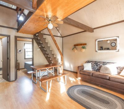 A cozy living room with a brown couch, a wooden dining table, a small staircase leading to a loft, and sunlight streaming in. There are rustic decorations, a ceiling fan, and a round rug on the wood floor.