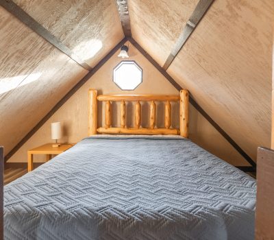 A cozy attic bedroom with sloped wooden ceilings, a rustic wooden bed frame, a blue quilted bedspread, a small side table, and a round window above the bed letting in light.