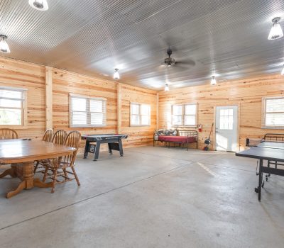 Spacious room with wood-paneled walls, a dining table with chairs, a foosball table, a ping pong table, and two daybeds under multiple windows and ceiling lights. The floor is concrete and the ceiling is metal.