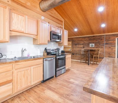A spacious kitchen with light wood cabinets, stainless steel appliances, and a long wooden countertop. The ceiling and walls are wood-paneled, and there is a small dining table in the background. The space is brightly lit.