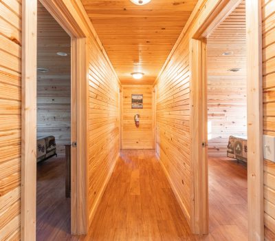 A bright hallway with wood-paneled walls, ceiling, and floor, leads to two bedrooms on either side. Natural light fills the space, and a picture and fire extinguisher are mounted at the end of the hall.