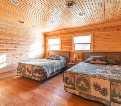 A cozy bedroom with wood-paneled walls and ceiling, featuring two beds with nature-themed quilts, a small nightstand with a lamp between them, and sunlight streaming through two windows above the beds.