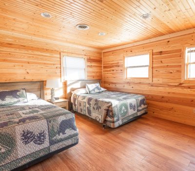 A cozy bedroom with wooden walls and ceiling, two beds with nature-themed quilts, matching pillows, a bedside table, and windows letting in natural light. The floor is polished wood, creating a warm, cabin-like atmosphere.