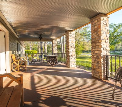 A spacious covered patio with wooden benches, a grill, and a table with chairs. Stone columns support the roof, and sunlight casts shadows on the concrete floor. Green trees and grass are visible in the background.