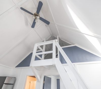 A white loft space with a high, slanted ceiling, a ceiling fan, and a staircase leading to an upper area. The walls and ceiling are painted white with blue-gray accents around the loft.