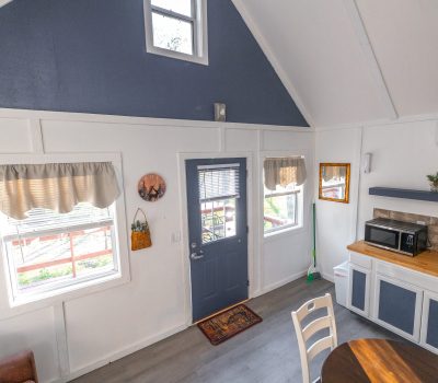 A bright, cozy kitchen and dining area with white walls, blue accents, large windows, a door with a window, countertop with microwave and sink, and a round table with chairs. Sunlight streams in, creating a welcoming feel.