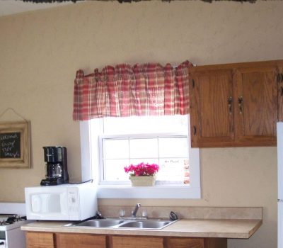 A kitchen featuring a sink with a cupboard underneath, a microwave, and a coffee maker on the counter to the left. There's a window with a red checkered valance and a potted plant on the windowsill. Above the sink, wooden cabinets are present. A chalkboard on the wall reads “EAT.”.