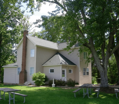 A tan two-story house with a brick chimney stands in a grassy area surrounded by tall leafy trees. Two picnic tables are placed on the lawn in front of the house, providing a serene outdoor setting. The sky above is clear and blue.